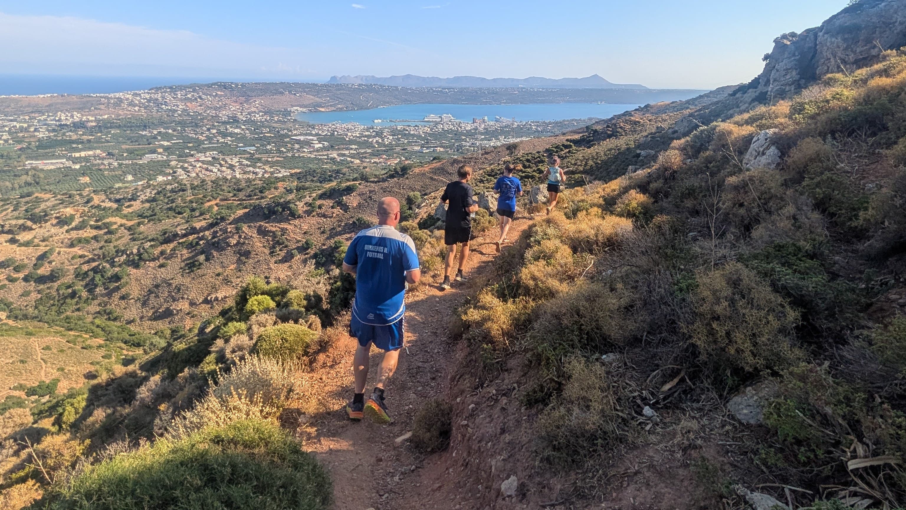 Four runners descending a mountain trail on the Malaxa range above Chania, with the bay and city below